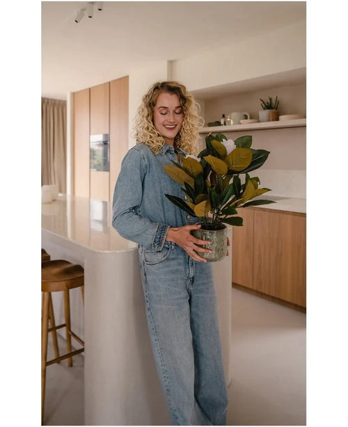 Una donna con i capelli biondi ricci, vestita con abiti in denim, sta sorridente in una cucina moderna con la pianta di magnolia artificiale in un vaso da 46 cm tra le mani, circondata da armadi e scaffali in legno: la scelta ideale per chi cerca piante a bassa manutenzione per migliorare i propri interni eleganti.