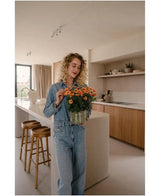 Una donna con i capelli biondi ricci, che indossa una camicia di jeans e jeans, si trova in una cucina moderna con una pianta artificiale Daisy UV di 31 cm tra le mani con fiori di colore arancione brillante. Accanto a lei ci sono due sgabelli di legno sull'isola della cucina.
