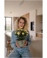 Una donna con i capelli biondi ricci, vestita con abiti in denim, siede in una cucina moderna con la Pianta Artificiale Mini Rose UV 35 cm gialla tra le mani e guarda dritto nella telecamera.