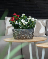 Una pianta artificiale Patio Rose 35 cm crema UV si trova su un tavolo rotondo in legno di fronte a un divano beige con cuscini fantasia.