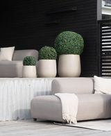 Quattro bossi artificiali in stile romano UV D40 cm, rotondi e beige, con arbusti verdi ben curati, sono posizionati su un patio moderno con sedie grigio chiaro, una coperta beige e uno sfondo in legno scuro per un look minimalista.