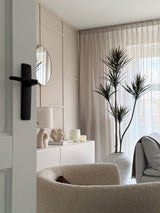 Un soggiorno moderno e minimalista con poltrona strutturata beige, pianta artificiale Dracaena Fragnans 180 cm, credenza bianca con decorazioni, specchio rotondo e tende trasparenti che filtrano la morbida luce del giorno attraverso la finestra.