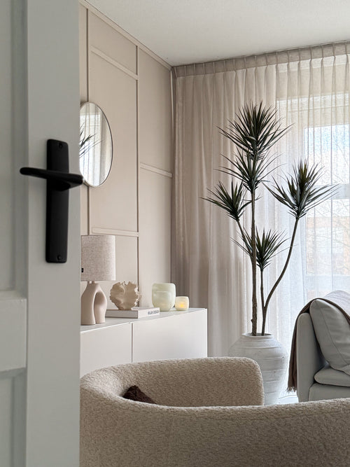 Un soggiorno moderno e minimalista con poltrona strutturata beige, pianta artificiale Dracaena Fragnans 180 cm, credenza bianca con decorazioni, specchio rotondo e tende trasparenti che filtrano la morbida luce del giorno attraverso la finestra.