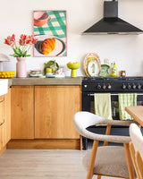 Una cucina luminosa con mobili in legno, fornello nero, asciugamani verdi, stoviglie colorate e l'immagine incorniciata di un dolce su un tavolo a scacchi acquista un aspetto fresco grazie al Fiore Artificiale Tulipano Parrocchetto Francese 76 cm rosso/giallo. Un'accogliente sedia bianca si trova accanto a un tavolo da pranzo in legno chiaro.