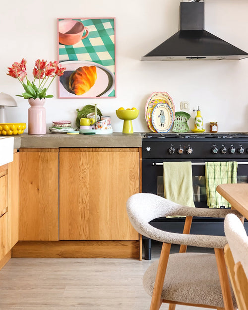 Una cucina luminosa con mobili in legno, fornello nero, asciugamani verdi, stoviglie colorate e l'immagine incorniciata di un dolce su un tavolo a scacchi acquista un aspetto fresco grazie al Fiore Artificiale Tulipano Parrocchetto Francese 76 cm rosso/giallo. Un'accogliente sedia bianca si trova accanto a un tavolo da pranzo in legno chiaro.