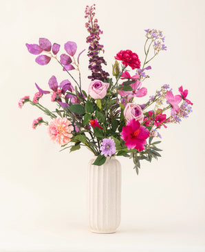 Un vaso strutturato bianco contiene il bouquet di fiori artificiali in seta Purple Dawn in tonalità rosa, viola e lavanda con fogliame verde, su uno sfondo chiaro e neutro.