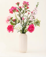 Un vaso testurizzato bianco mostra il bouquet di fiori artificiali in seta Blush Breeze - fiori di seta rosa, bianchi e viola chiaro con foglie verdi - su uno sfondo chiaro.