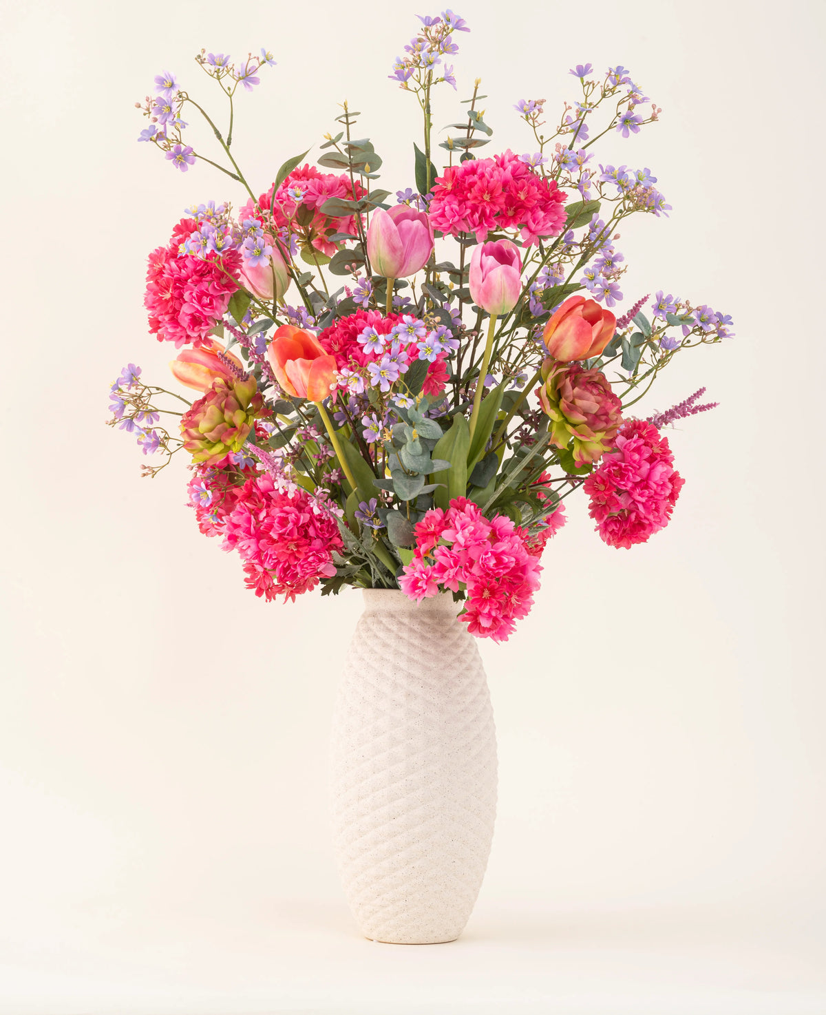 Un vaso con struttura bianca e il bouquet di fiori artificiali in seta Soft Spring con fiori rosa brillante, tulipani arancioni e fiori viola-blu portano la primavera nella tua casa su uno sfondo chiaro e semplice.