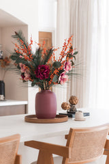 Il bouquet di fiori artificiali Silk Cozy Winter in un vaso color malva, con fiori rosa, bacche arancioni e rami di pino, adorna un vassoio rotondo in legno su un tavolo bianco per un look autunno/inverno accogliente e moderno con sedie in legno e tende beige.