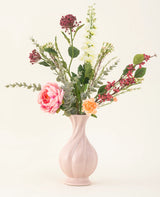 Il bouquet di fiori artificiali in seta Pretty Petite, in un vaso rosa pastello, contiene fiori rosa e arancioni fatti a mano, fiori bianchi, foglie verdi e bacche rosse - perfetti per un interno sostenibile su uno sfondo chiaro e semplice.