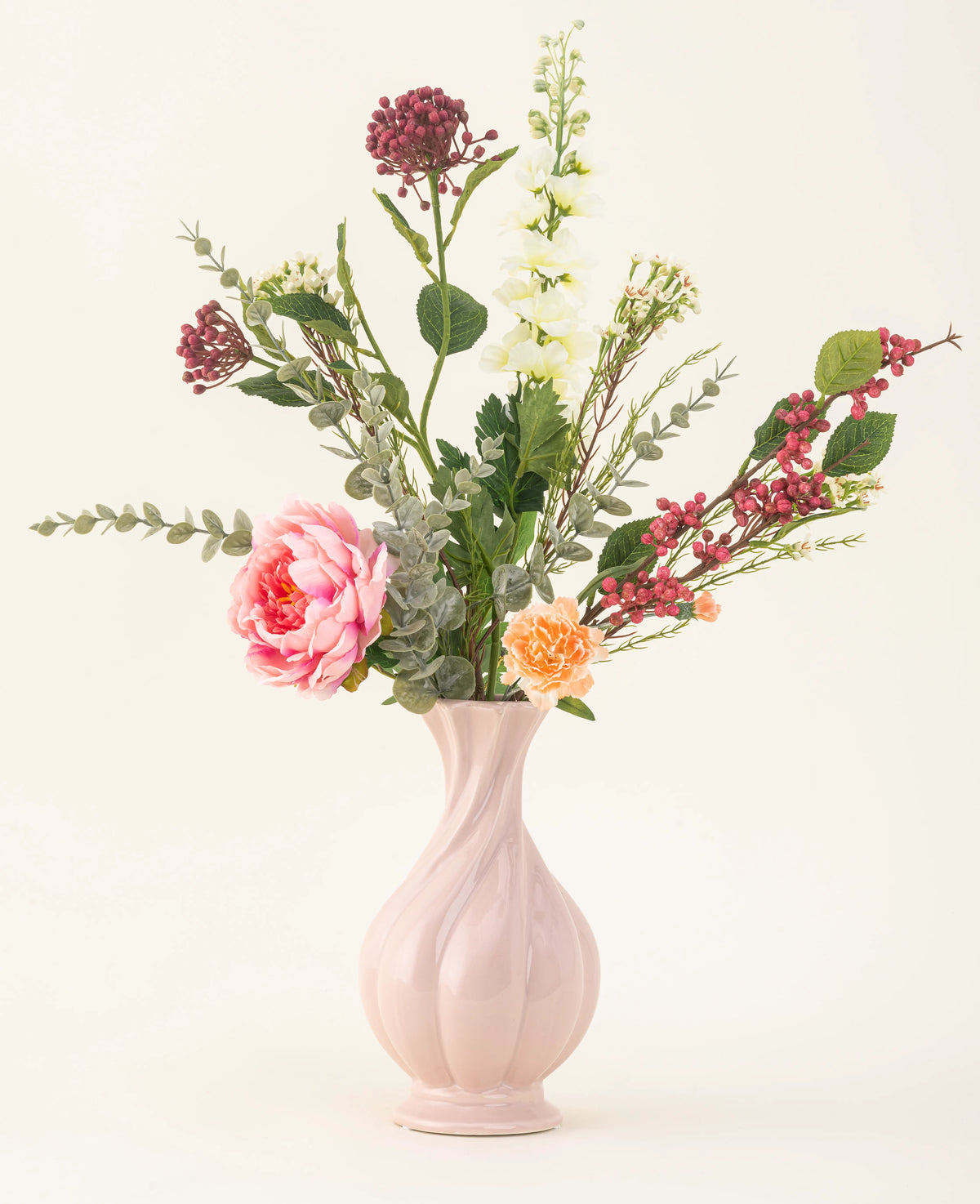 Il bouquet di fiori artificiali in seta Pretty Petite, in un vaso rosa pastello, contiene fiori rosa e arancioni fatti a mano, fiori bianchi, foglie verdi e bacche rosse - perfetti per un interno sostenibile su uno sfondo chiaro e semplice.