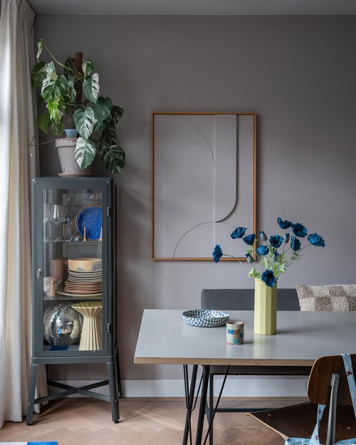 La moderna sala da pranzo è dotata di una vetrina in vetro grigio in cui sono esposte stoviglie e piante, decorazioni murali minimaliste e un tavolo su cui poggia il fiore artificiale Papavero 73 cm blu scuro in un vaso giallo. Lo spazio è completato da un eclettico mix di sedie, il tutto su uno sfondo di pareti grigie e un pavimento in legno.