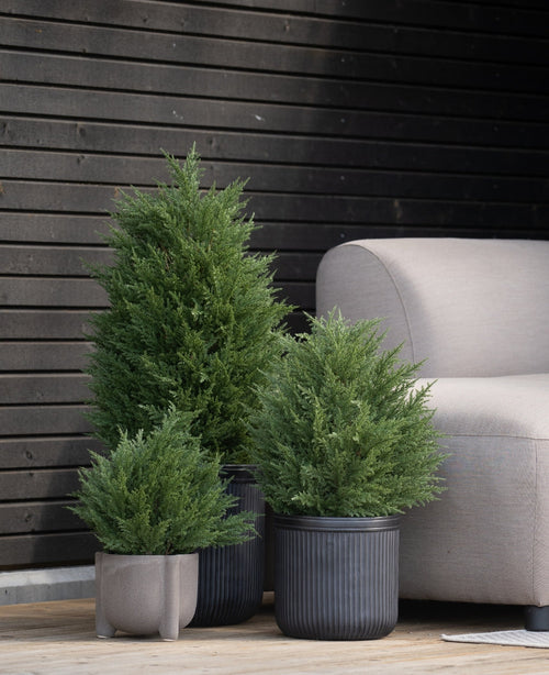 Le conifere artificiali Artificial Conifer UV 40 cm sono posizionate su una terrazza in legno accanto a un divano da esterno grigio chiaro, contro una parete moderna con pannelli in legno scuro.
