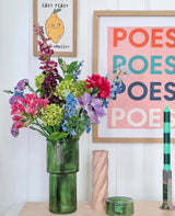 Un vaso di vetro verde contiene un bouquet di fiori artificiali di seta Blossom Breeze sul tavolo. Dietro sono appese delle foto incorniciate: una con un limone e "EASY PEASY", un'altra con "POESY" - che aggiungono ulteriore fascino. Nelle vicinanze ci sono una candela verde e una candela a spirale rosa di Blossom Breeze.