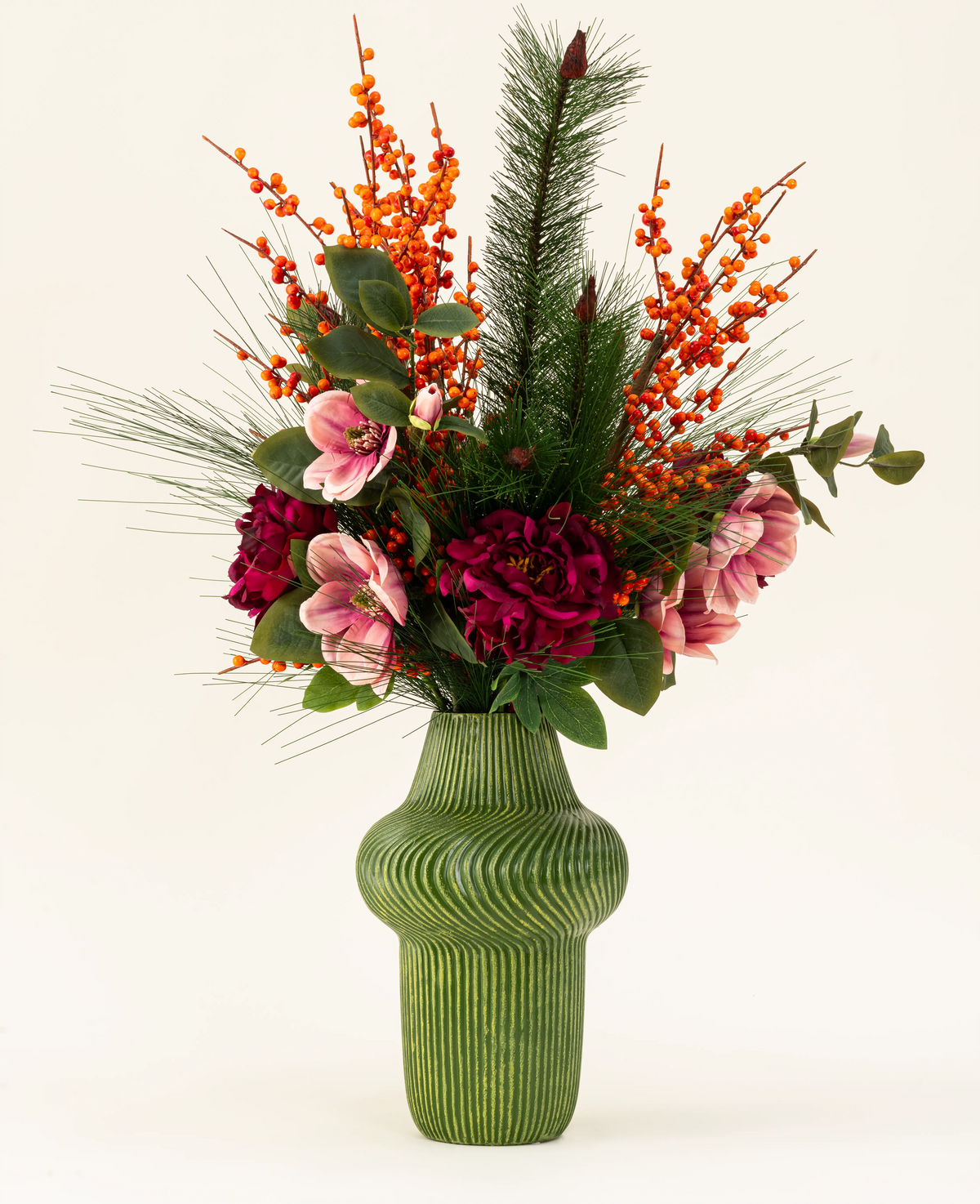 Il bouquet di fiori artificiali Silk Cozy Winter presenta un vaso verde a coste con fiori rosso scuro e rosa, bacche arancioni, rami di pino e foglie verdi - perfetto come decorazione autunno-inverno su uno sfondo chiaro.