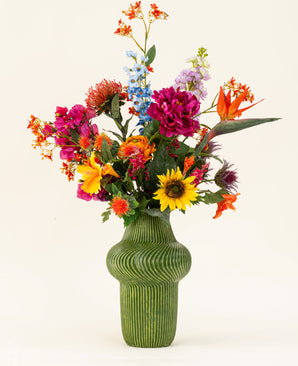 Un vaso strutturato verde mostra il bouquet di fiori artificiali in seta Colourful Spirit, con girasoli, peonie rosa, fiori arancioni e viola e una vegetazione lussureggiante su uno sfondo chiaro e semplice.
