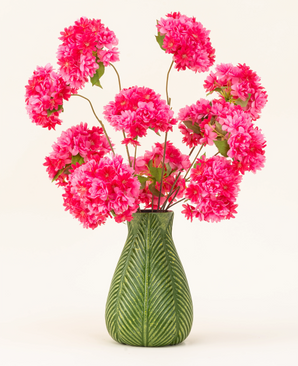 Un vaso verde con motivo a foglie mostra il bouquet di fiori di seta artificiale Cherry Blossom Pink - soffici fiori di ciliegio di seta rosa brillante con foglie verdi - su uno sfondo chiaro e semplice.