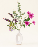 Disposto in un moderno vaso bianco, il bouquet di fiori artificiali in seta Lavender Skies presenta fiori artificiali rosa e viola, accenti gialli e fogliame verde lussureggiante: bellissimo su uno sfondo chiaro e semplice.