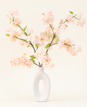 Un vaso in ceramica bianca con un'ampia apertura ovale mostra diversi bouquet di fiori artificiali in seta a foglie verdi Rami di fiori di ciliegio rosa chiaro su un semplice sfondo chiaro.