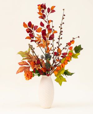 Un vaso strutturato bianco mette in mostra il bouquet di fiori artificiali in seta con fascino di castagna, caratterizzato da foglie autunnali, fiori d'arancio e fogliame verde: un'elegante composizione artificiale su uno sfondo chiaro e semplice.