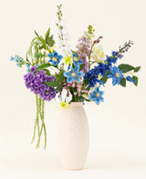 Un vaso strutturato bianco contiene il bouquet di fiori artificiali in seta Midnight Valley, caratterizzato da vivaci fiori Real Touch blu, viola e gialli con diverse foglie verdi su uno sfondo chiaro.