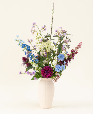 Un vaso bianco con il bouquet di fiori artificiali in seta Purple Rain mostra vivaci fiori artificiali viola e bianchi e foglie verdi, un esempio di artigianato olandese su uno sfondo chiaro.