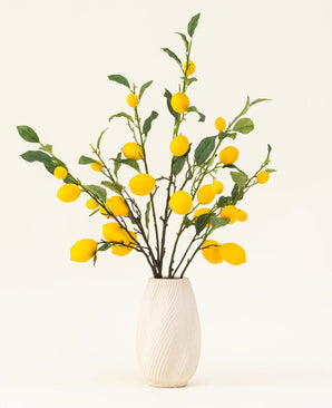 Un vaso bianco testurizzato mostra il bouquet di fiori artificiali in seta Lemons, con foglie verdi e vivaci limoni gialli su uno sfondo chiaro.