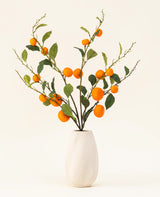 Il bouquet di fiori artificiali in seta Mandarino ha un vaso bianco strutturato e rami di foglie verdi e mandarini artificiali di colore arancione vibrante, che aggiungono un tocco vivace su uno sfondo chiaro e semplice.