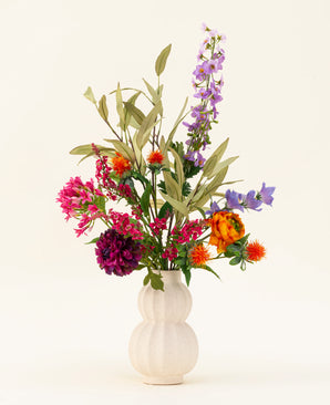 Un vaso bianco strutturato mette in mostra il bouquet di fiori artificiali in seta colorata Svenski, caratterizzato da vivaci fiori artificiali viola, arancioni, rosa e blu con foglie verdi che illuminano qualsiasi spazio su un semplice sfondo chiaro.
