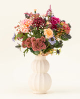 In un vaso di ceramica bianca e ricurva si trova il bouquet di fiori artificiali in seta Pretty Pastel - un mix colorato di fiori sostenibili come rose rosa, dalie, fiori viola e arancioni e vegetazione - splendidamente disposti su uno sfondo chiaro.