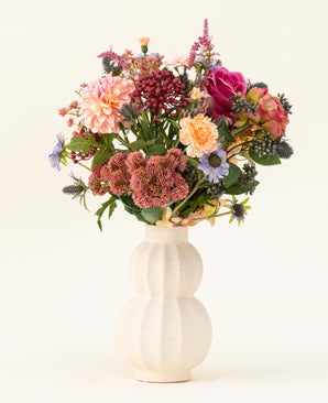 In un vaso di ceramica bianca e ricurva si trova il bouquet di fiori artificiali in seta Pretty Pastel - un mix colorato di fiori sostenibili come rose rosa, dalie, fiori viola e arancioni e vegetazione - splendidamente disposti su uno sfondo chiaro.