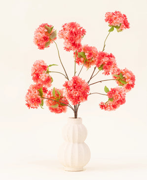 Il bouquet di fiori artificiali in seta Cherry Blossom Coral presenta vivaci fiori di ciliegio artificiali rosa e corallo con foglie verdi, elegantemente disposti in un vaso scolpito bianco su uno sfondo chiaro.