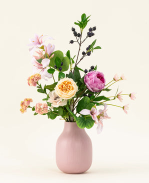 Il bouquet di fiori artificiali in seta Touch of Cloudlight, presentato in un vaso di ceramica rosa, contiene peonie rosa, rose gialle, piccoli fiori, verde e rami con bacche scure su uno sfondo chiaro.