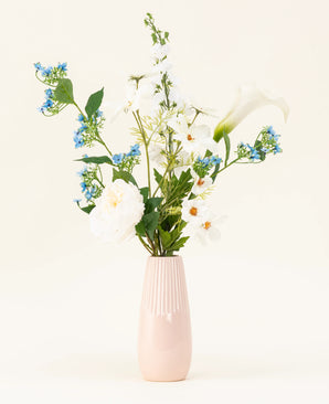 Il vaso rosa chiaro contiene il bouquet di fiori artificiali in seta Whispers of White, con fiori bianchi, fiori blu e fogliame verde su uno sfondo semplice e chiaro.