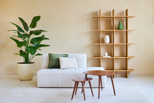 Soggiorno minimalista con divano bianco, due cuscini, tavolini impilabili in legno e una grande pianta artificiale Strelitzia Real Touch Deluxe 180 cm per un'atmosfera tropicale. Una moderna libreria aperta in legno con vasi si trova contro il muro beige.