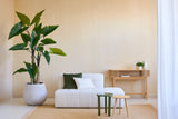 Un soggiorno minimalista con la pianta artificiale Calocasia 210 cm, un divano strutturato bianco con cuscini verdi e bianchi, due tavolini laterali, un tappeto beige e una consolle in legno contro una parete beige chiaro.