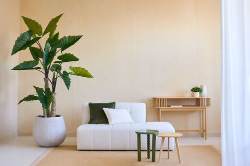 Un soggiorno minimalista con la pianta artificiale Calocasia 210 cm, un divano strutturato bianco con cuscini verdi e bianchi, due tavolini laterali, un tappeto beige e una consolle in legno contro una parete beige chiaro.
