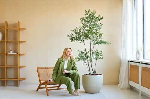 Una donna vestita di verde sorride mentre si siede su una sedia di legno accanto alla pianta artificiale Ruscus da 180 cm in una stanza moderna e luminosa con pareti beige, grandi finestre e arredamento minimalista.