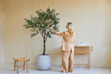Una donna in abiti larghi beige sta accanto a un olivo artificiale di 200 cm e tocca delicatamente le foglie. Nelle vicinanze, uno sgabello in legno e una consolle con libri e un diffusore sono appoggiati alla parete beige.