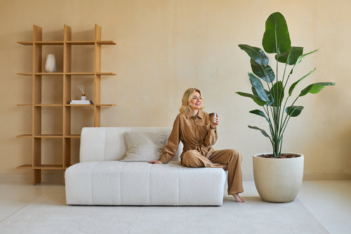 Una donna vestita di marrone è seduta su un divano bianco e sorride con una tazza in mano. Accanto a lei c'è la Pianta Artificiale Strelitzia 180 cm real touch, che porta una vegetazione lussureggiante nel moderno soggiorno dai toni beige con un arredamento minimal.