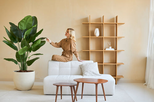 Una donna con una tuta marrone si siede sullo schienale di un divano bianco e tocca la foglia di una pianta artificiale Strelitzia Real Touch Deluxe da 180 cm in un soggiorno minimalista con tavoli e scaffali in legno.