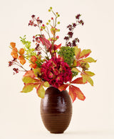 In un vaso marrone a coste si trova il bouquet "Fiori artificiali di seta Ambra calda", con fiori e foglie vivaci di colore rosso, arancione e verde su lunghi steli, su uno sfondo chiaro e semplice.
