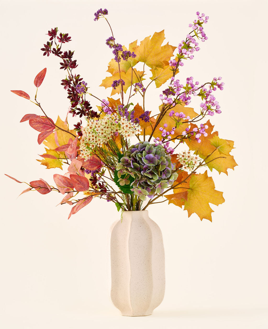 Il bouquet di fiori artificiali di seta "Wild Amethyst", con foglie autunnali, fiori viola e bianchi e ortensie verdi, è in un vaso di ceramica bianca su uno sfondo semplice e chiaro.