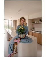 Una donna dai capelli biondi e ricci, che indossa jeans e calzini a righe, siede sorridente sul bancone della cucina mentre tiene in mano la pianta Artificiale Gardenia UV 39 cm – una pianta in vaso con fiori gialli. Sullo sfondo puoi vedere una cucina moderna con mobili e scaffali in legno.
