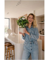 Una donna sorridente in abito jeans tiene in mano la pianta artificiale bianca Artificial Gardenia UV 39 cm in una cucina moderna con sgabelli in legno, piani di lavoro bianchi e decorazioni neutre.