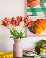 Su un tavolo con stoviglie colorate si trova un vaso rosa contenente il fiore artificiale Tulipano Parrocchetto Francese 76 cm rosso/giallo, mentre sullo sfondo si vede in parte un dipinto incorniciato di un croissant e una tazza.