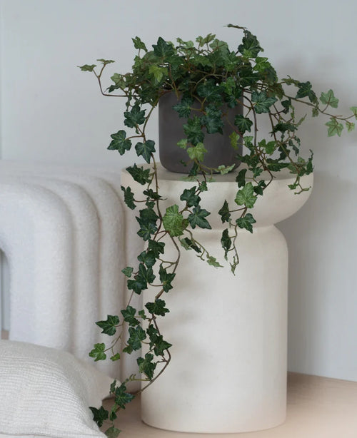 Un'edera artificiale Deluxe Green, un'edera pendente di 90 cm in un vaso grigio, si trova su una base moderna, bianca e scultorea accanto a una coperta bianca piegata e un cuscino strutturato bianco.