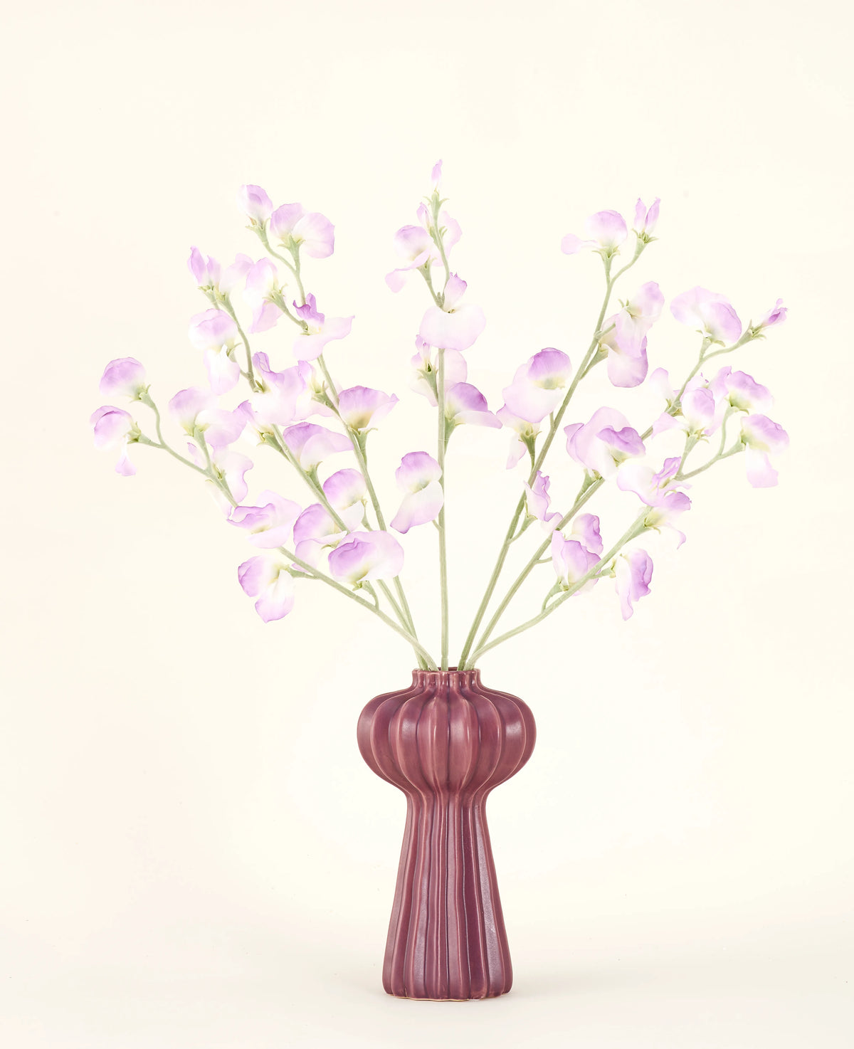 Il bouquet di fiori artificiali in seta Pea lilla presenta lunghi steli di delicati fiori viola chiaro e foglie verdi in un vaso di ceramica color malva con creste verticali, su uno sfondo semplice e bianco sporco.