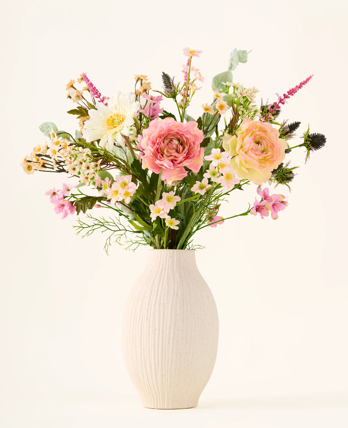 In un vaso di ceramica bianca si trova il bouquet di fiori di seta artificiali "Backyard Blossom", costituito da una bellissima composizione di rose rosa, margherite bianche e piccoli fiori gialli, realizzati con fiori di seta, integrati con rigogliose foglie verdi su uno sfondo chiaro.