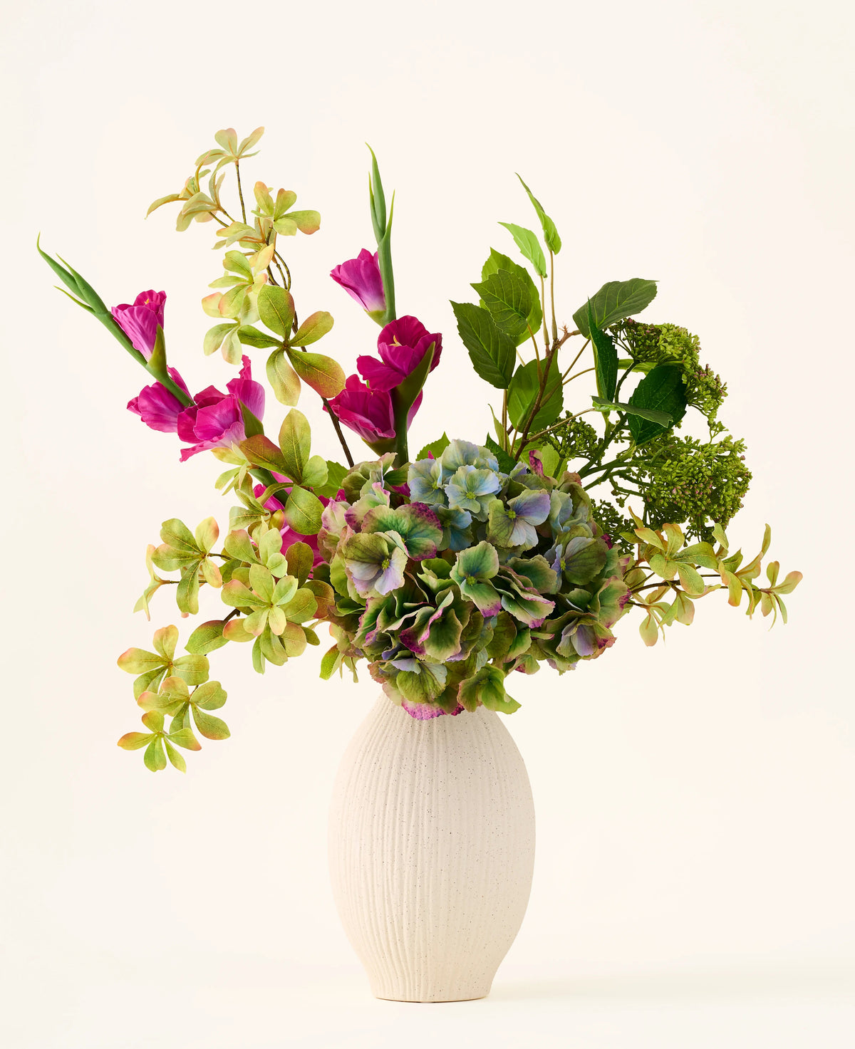 Un vaso strutturato bianco contiene un bouquet di ortensie verdi, rami frondosi e fiori di seta magenta brillante su uno sfondo chiaro e semplice, creando un sorprendente bouquet di fiori di seta artificiali "Cottage Garden" che aggiungerà un fascino duraturo a qualsiasi spazio.